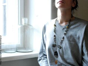 Detoxdesign Na nastrojowym zdjęciu kobieta siedząca przy oknie w pięknym oknie nosi zachwycający długi naszyjnik SHIRAZU05 z pięknych kamieni.