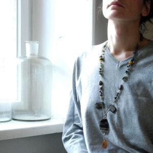 Na nastrojowym zdjęciu kobieta siedząca przy oknie w pięknym oknie nosi zachwycający długi naszyjnik SHIRAZU05 z pięknych kamieni.